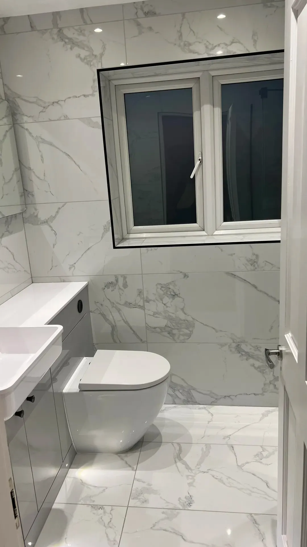 Basin and back-to-wall toilet under a window, fully tiled in marble effect